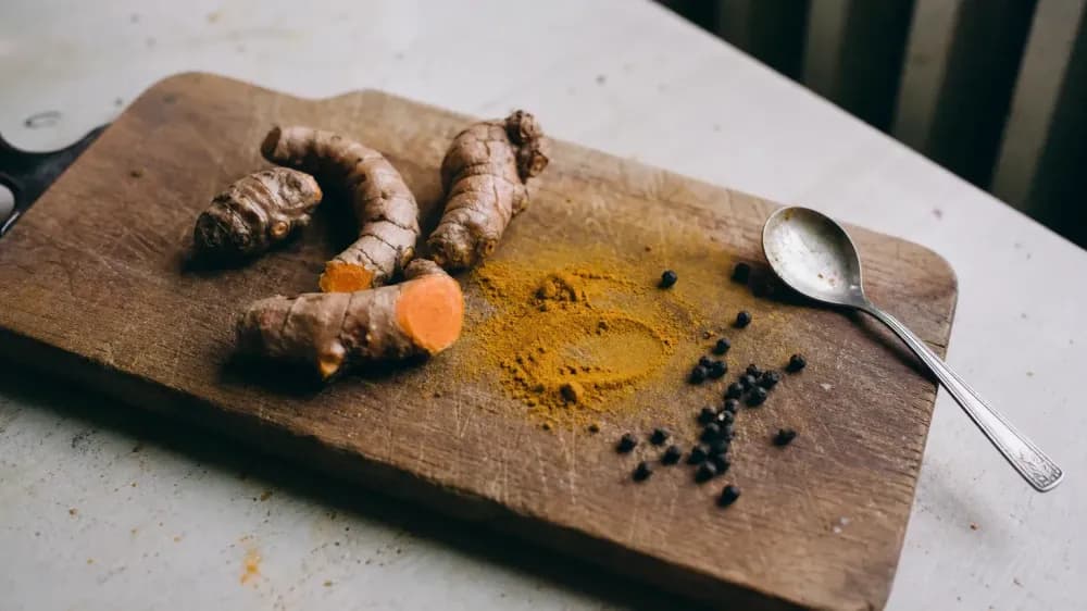 Fresh turmeric rhizomes, a spill of ground turmeric and loose black peppercorns on a scratched wooden board in daylight.