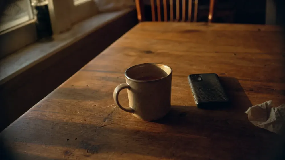 A stoneware mug of tea left untouched on a scratched wooden table beside a phone lying face down, in weak evening light from a window at the left, with nobody in the room.