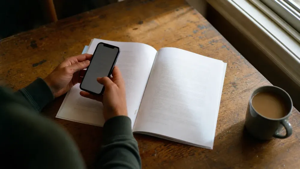 Two hands holding a phone above an open stapled printout on a wooden table by a window, the text on both the screen and the pages too shallow to read.