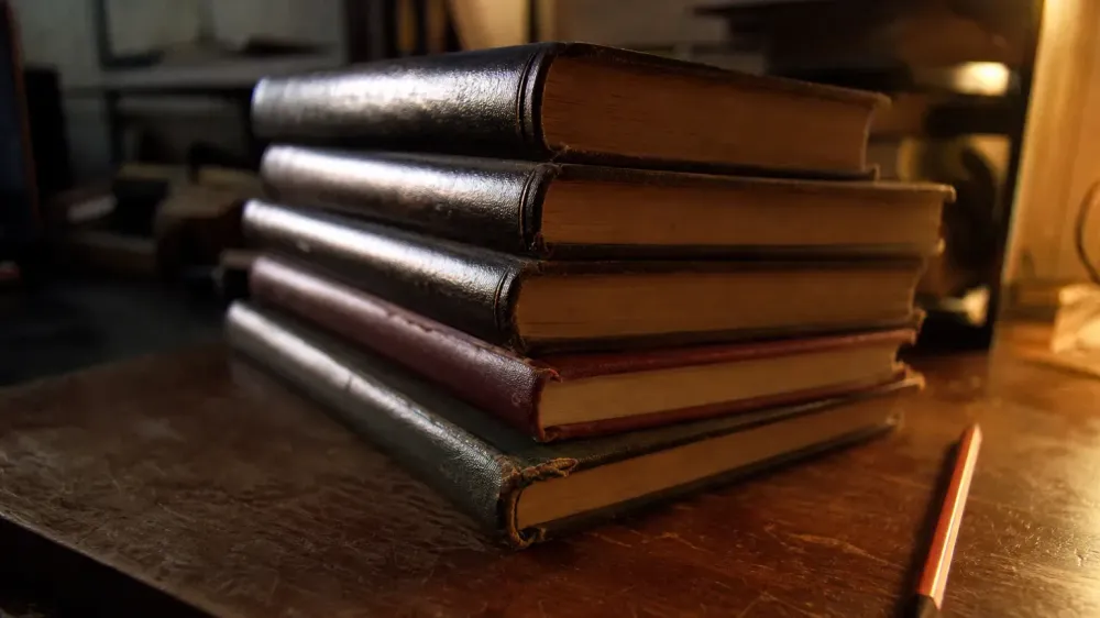 A stack of worn hardback journal volumes on a desk under a low lamp, spines angled away from the camera so no title can be read.