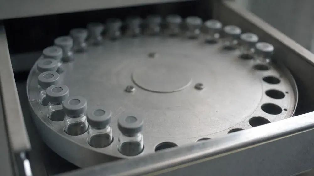 A row of small clear glass autosampler vials with plain septum caps seated in a metal carousel tray inside an open instrument drawer, lit by flat laboratory daylight.