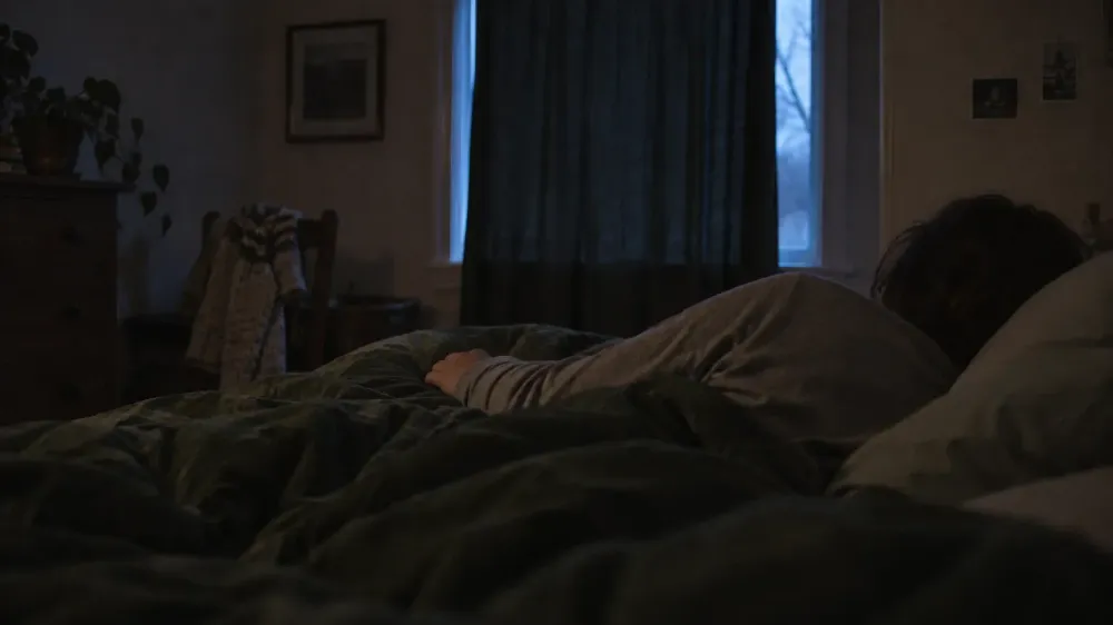 A person lying awake on their side in a dark bedroom, seen from behind, with faint early light at the window and rumpled covers.