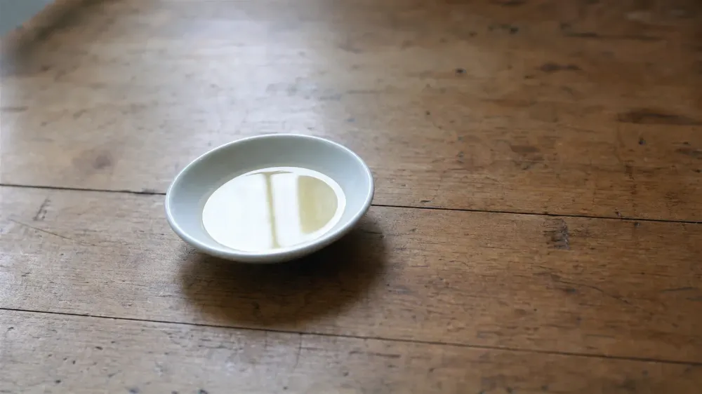 A small white ceramic dish holding a shallow pool of pale golden oil on a worn wooden kitchen table, photographed in soft morning window light with the background falling out of focus.
