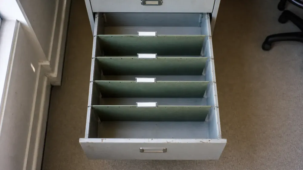 An open metal filing drawer holding four empty hanging folders with nothing filed in them, lit by plain daylight from a window at the side.