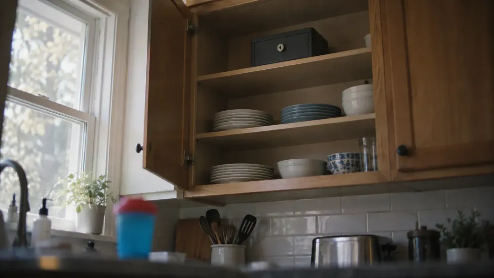 A small locked metal box on the top shelf of an open upper kitchen cabinet, above everyday dishes and well out of a child's reach.