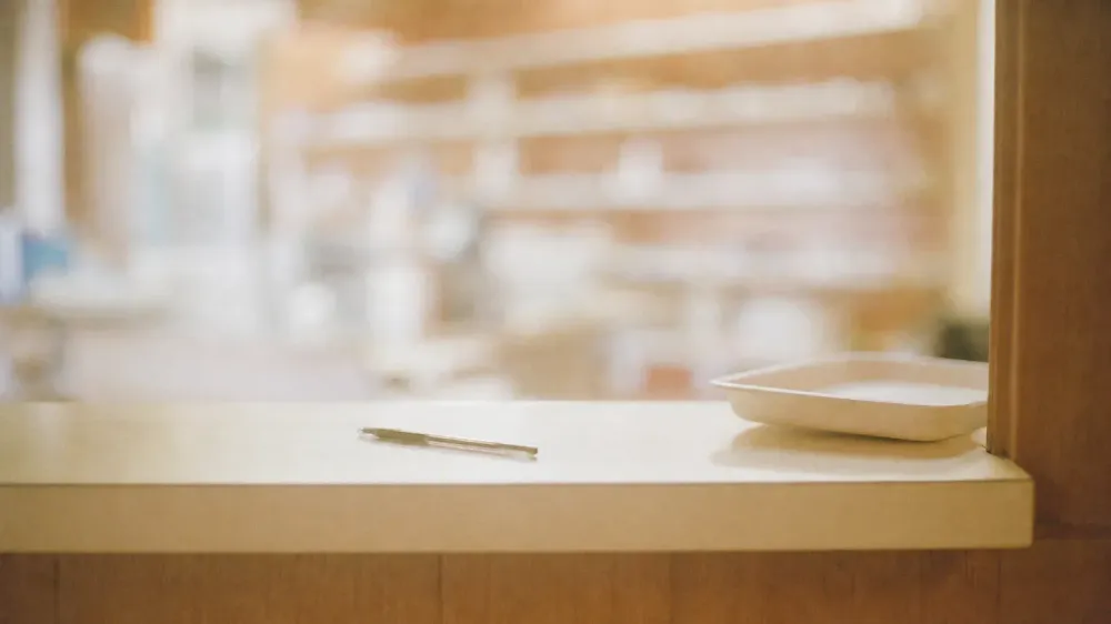 A quiet community pharmacy consultation counter photographed from the customer's side, with a clean empty counter, a pen, and heavily blurred shelving behind it.
