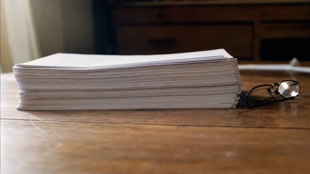 A thick stack of loose paper resting on a worn wooden desk, photographed almost edge on so only the page edges are in view, with a pair of reading glasses folded beside it.