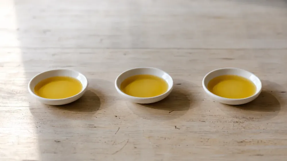 Three small identical white ceramic dishes in a row on a pale wooden bench, each holding a shallow pool of golden hemp oil, indistinguishable from one another in daylight.