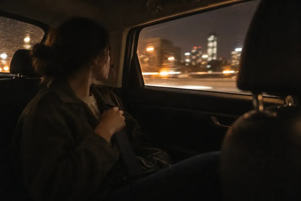 A passenger in the back seat of a car at night fastening a seatbelt, with blurred city lights passing through the side window.