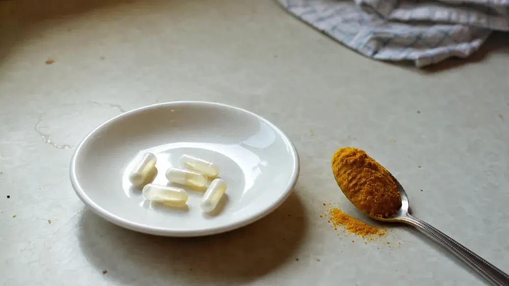 A plain white saucer holding a few loose unmarked capsules beside a teaspoon of bright ground turmeric on a pale kitchen counter.