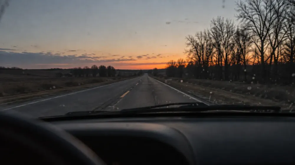 An empty two-lane road at dusk seen through a car windshield, wipers at rest, the dashboard a dark silhouette against a pale sky.