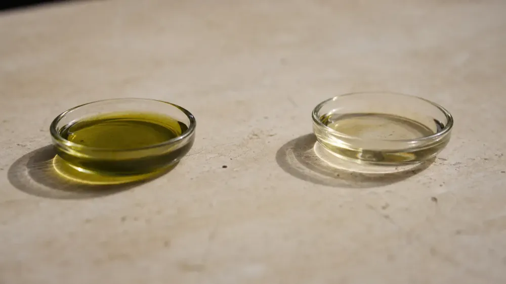 Two small shallow glass dishes on a pale stone surface, one holding a greenish oil and the other a clear pale gold oil, set apart from each other in hard side light.