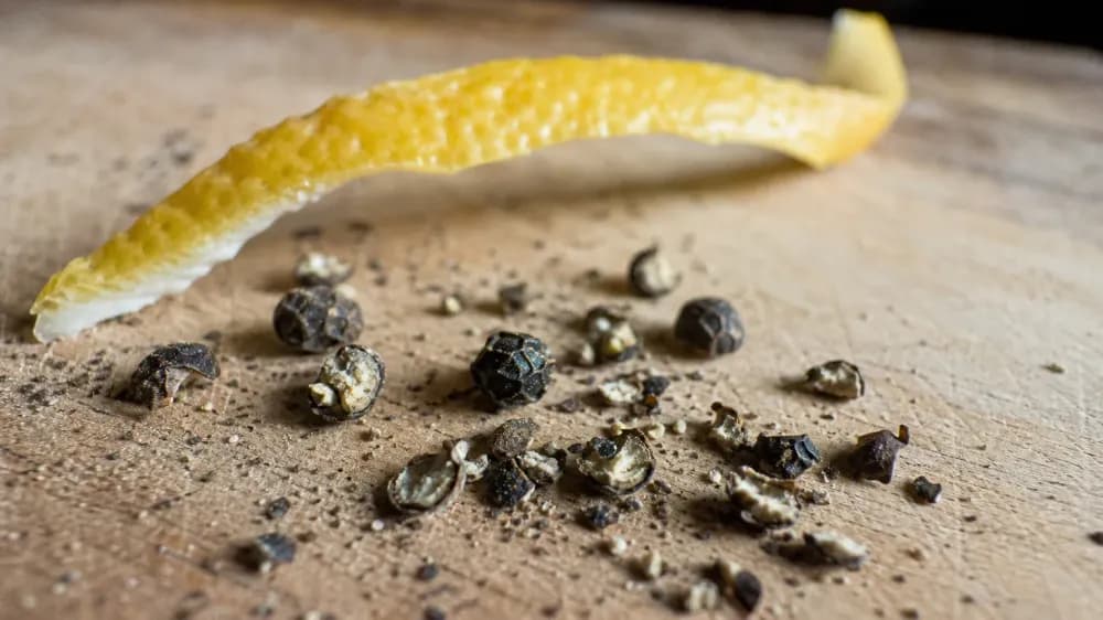 Macro photograph of cracked black peppercorns and a curl of lemon peel on a worn wooden board in daylight.