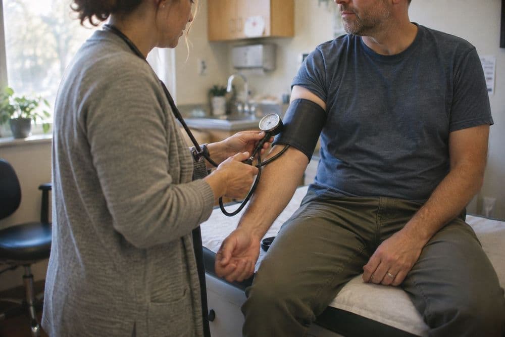 A clinician fitting a blood pressure cuff on the upper arm of an adult patient seated on an exam table in a small, ordinary clinic room.