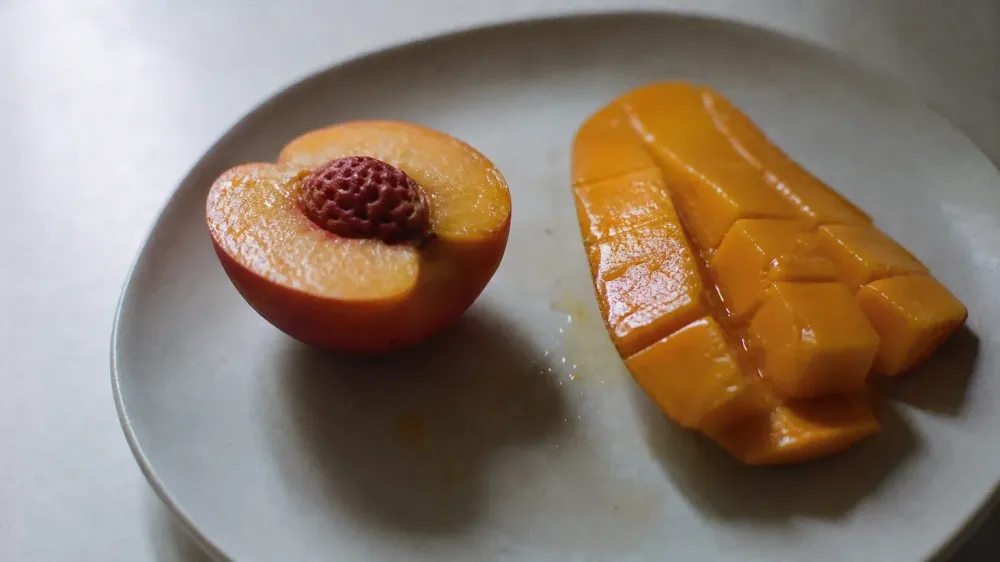 A halved ripe peach and a cut cheek of mango on a pale ceramic plate in daylight.