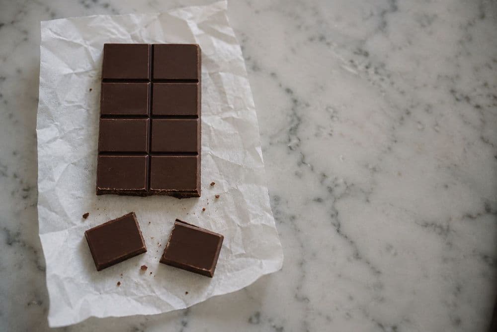An unbranded dark chocolate bar scored into a grid of equal squares, with two squares snapped off along the score lines and lying beside it on parchment paper on a marble counter.