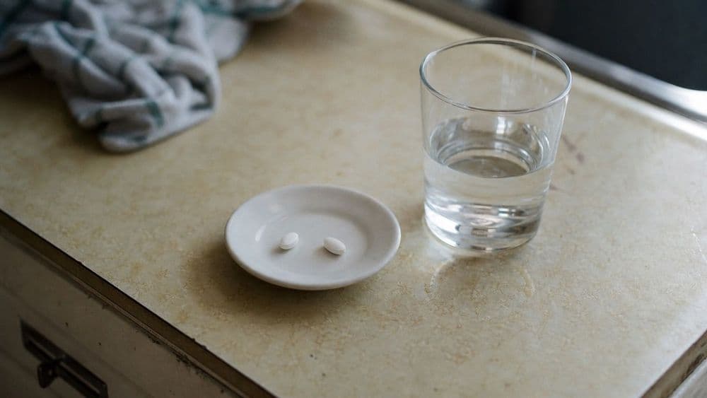 Two plain unmarked white tablets on a small saucer beside a glass of water on a worn kitchen counter in morning light.