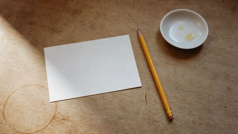 A blank white index card and a sharpened pencil on a worn wooden table beside a small white dish holding a few drops of pale golden oil.