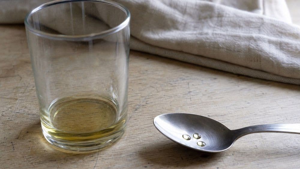 A small clear glass holding a shallow pool of pale gold oil on a worn wooden counter, with a plain steel teaspoon beside it holding only three drops.