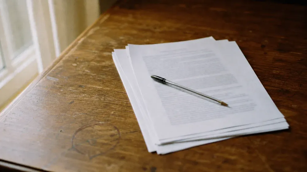 A loose stack of printed report pages on a worn wooden desk beside a window, a ballpoint pen lying across the top page, photographed at a low angle so the printed type reads only as soft grey lines.