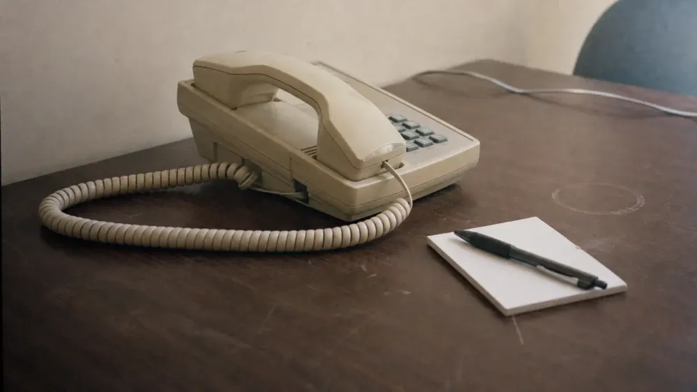 A corded desk telephone on an otherwise bare office desk, the handset resting slightly askew in its cradle beside a blank notepad.