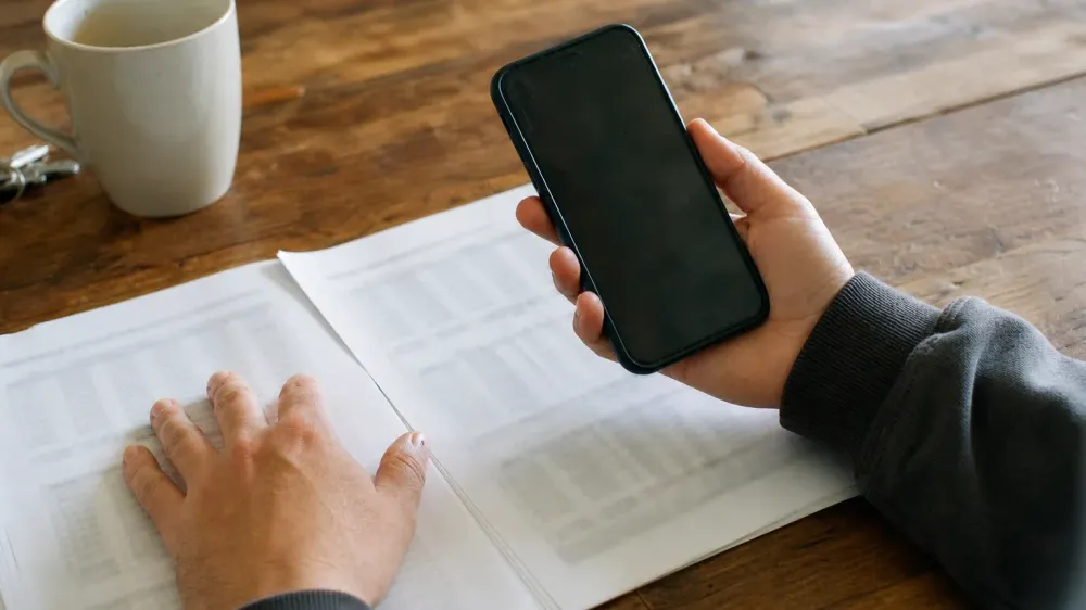 A person's hands holding a phone above a printed multi-page laboratory report on a kitchen table, comparing the two.