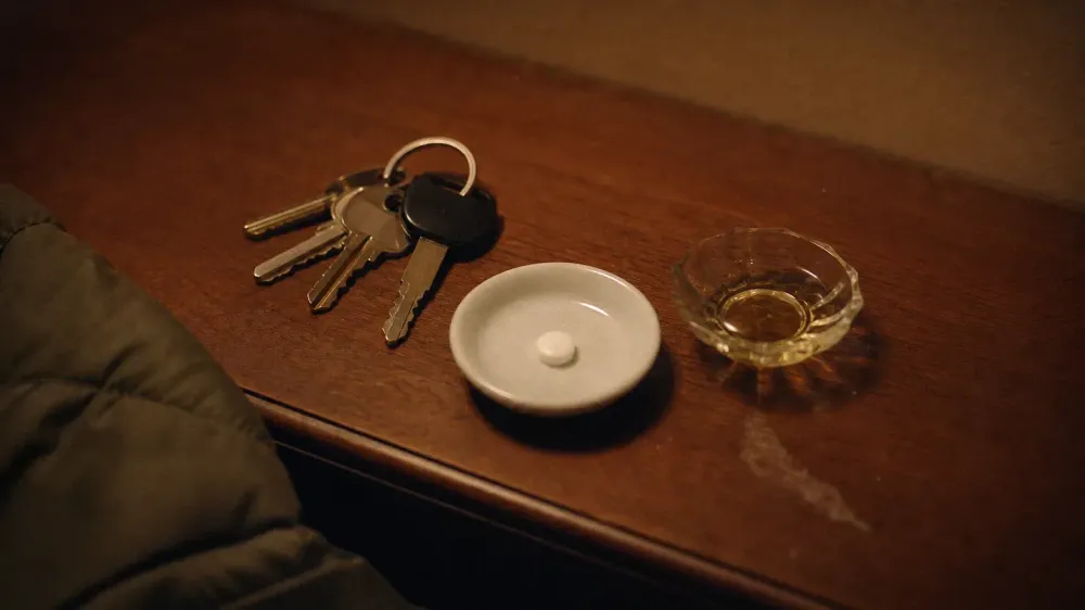 House and car keys on a wooden entry table in low evening light, beside a white saucer holding one unmarked round white tablet and a small dish of pale golden oil.