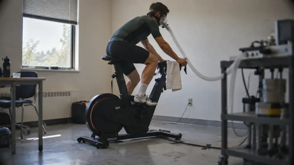 A cyclist on a stationary lab bike wearing a breathing mask connected to a gas-analysis hose, photographed from the side in a plain exercise physiology lab.