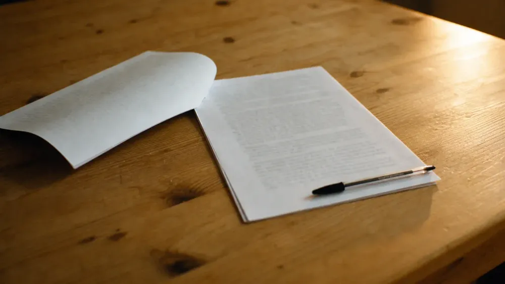 A stapled multi-page laboratory report lying open on a wooden kitchen table beside a ballpoint pen, the body type too shallow to read.