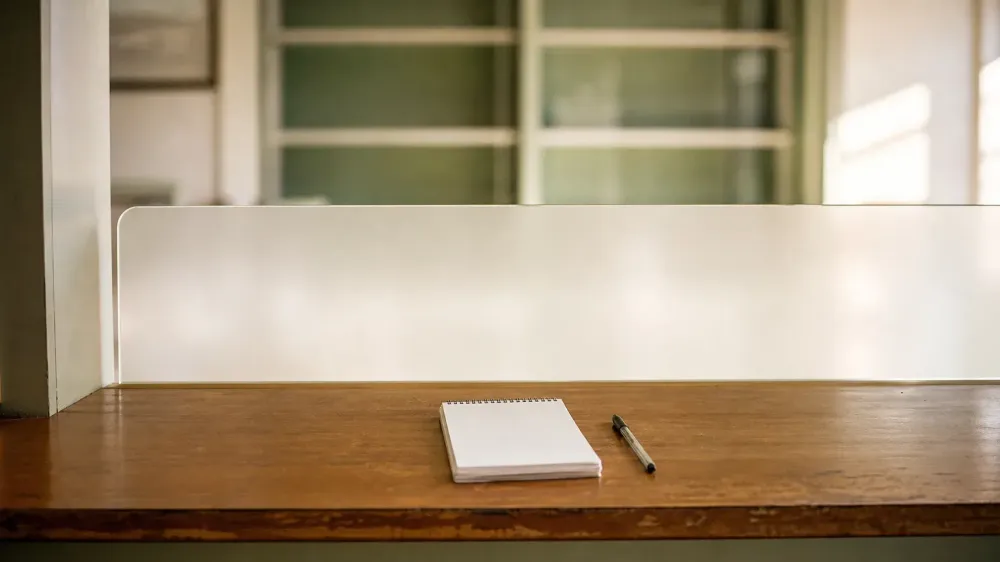 An empty pharmacy consultation counter in warm daylight, with a blank notepad and a pen on the wooden surface and a frosted glass partition behind it.