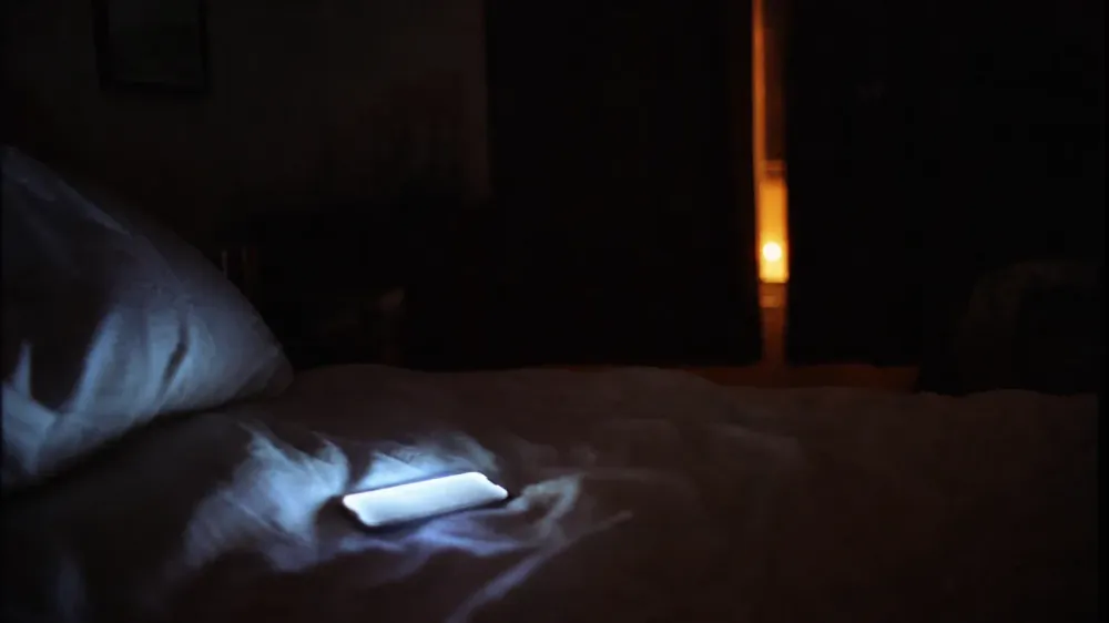 A dark bedroom at night with the glow of a phone screen lighting a rumpled duvet, the screen content out of focus and unreadable.