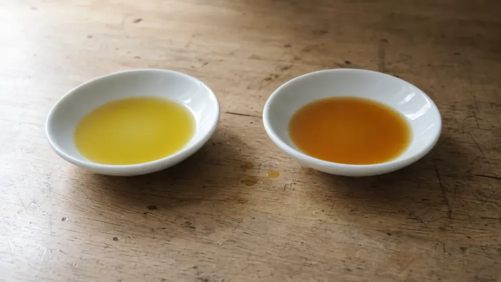 Two small white dishes side by side on a wooden table, one holding pale gold oil and the other holding a deeper amber oil, in natural window light.