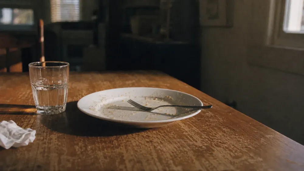 A used dinner plate and fork left on a scuffed wooden table in low evening light from a single window, with a half-empty water glass beside them and nobody in the room.