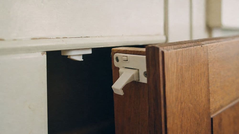 A white plastic child-safety latch fitted inside a wooden kitchen cabinet door, the door held slightly ajar so the latch arm and its catch are visible.