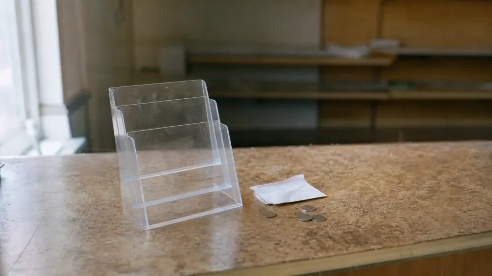 A worn laminate shop counter photographed close up, with a small empty acrylic display stand, a folded paper receipt and a few coins, and out-of-focus shelving behind. No packaging is visible.
