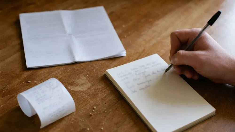 A hand writing a short note with a ballpoint pen on a paper pad, beside a folded printout and a curled paper receipt on a kitchen table.