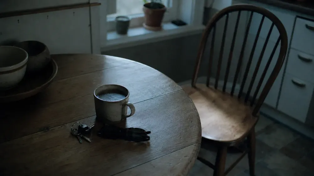 A kitchen table in early morning daylight with a cup of coffee, car keys and sunglasses beside an empty chair.