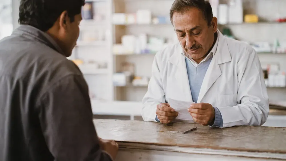 A person at a pharmacy counter handing a handwritten list of everything they take across to a pharmacist.