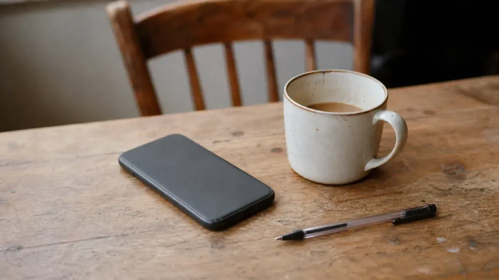 A phone lying face down on a worn wooden kitchen table beside a half-drunk mug of tea in morning light, with nobody in the frame.