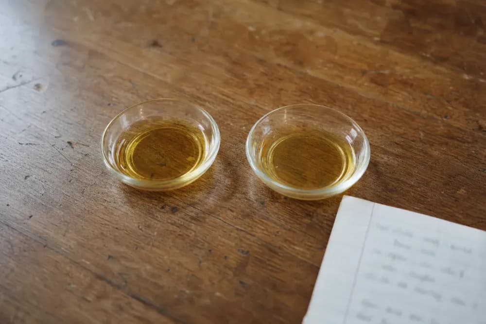 Two identical small glass dishes side by side on a worn wooden table, each holding a shallow pool of amber oil, visually indistinguishable from one another.