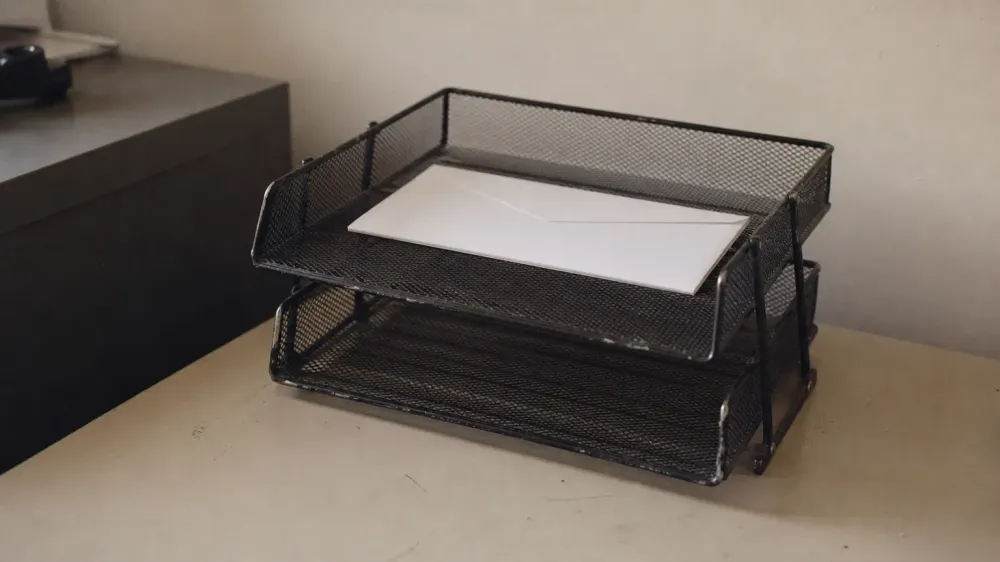 Two stacked wire desk trays on a plain office desk, the upper tray holding a single unmarked white envelope and the lower tray empty.