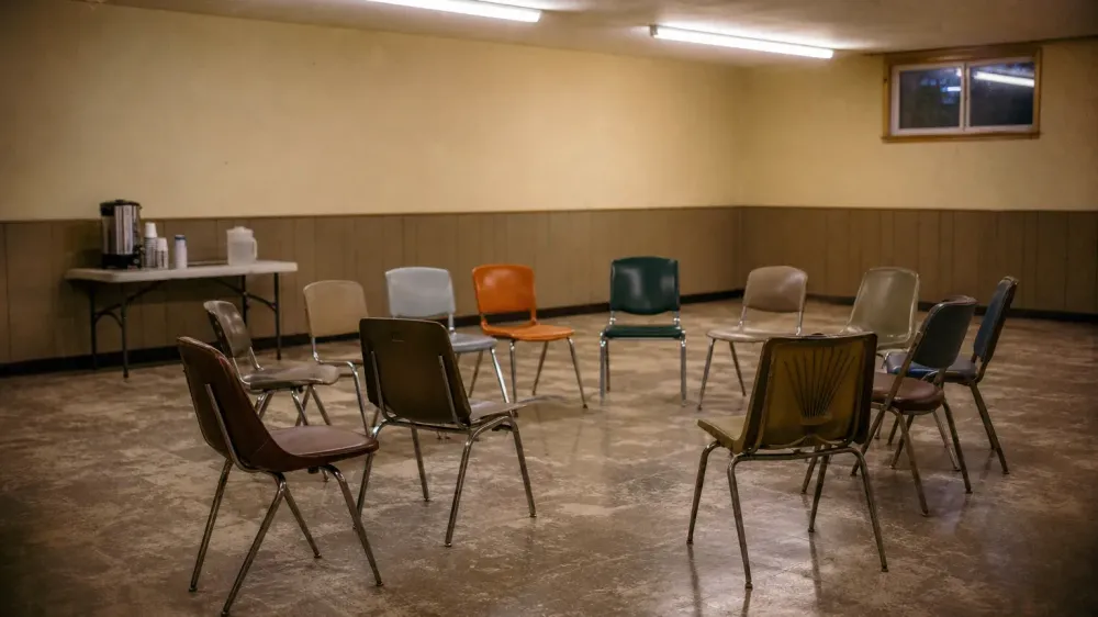 An empty community meeting room in the evening, stacking chairs arranged in a loose circle, a coffee urn on a folding table against the wall.