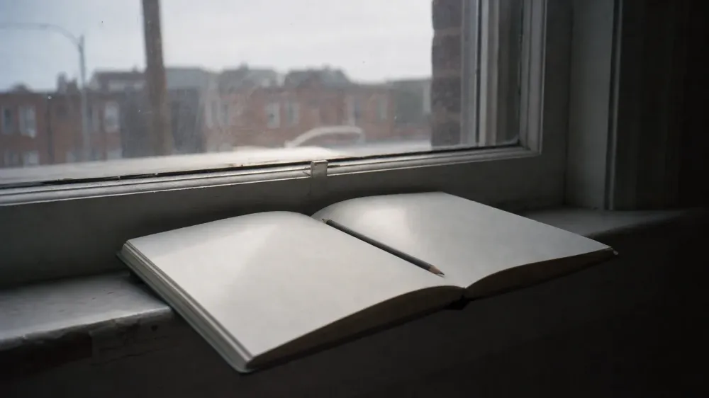 An open notebook with a pencil resting in the gutter, left on a wide windowsill in flat overcast daylight, with the page angled away so no writing is legible and nobody in the room.