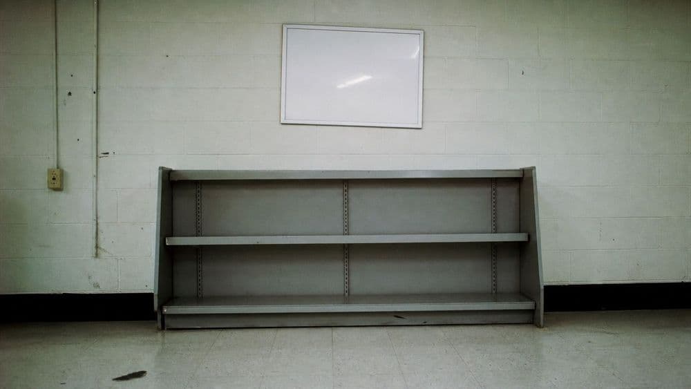 A blank white poster frame mounted on the wall of a small store, above an empty metal shelf, lit by overhead fluorescent light.
