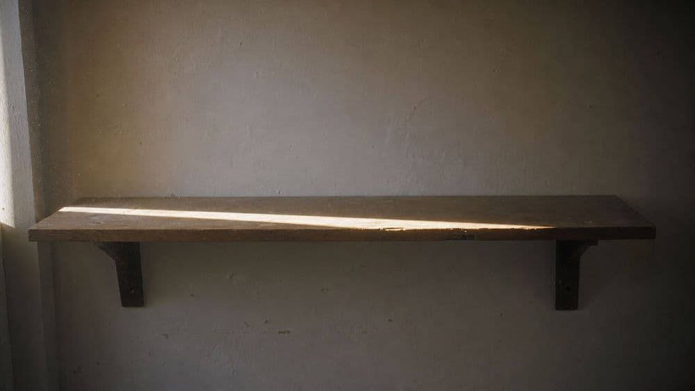 Late afternoon sunlight falling through a window onto a plain wooden shelf, one bright band of light across the wood and the rest of the shelf in shadow.