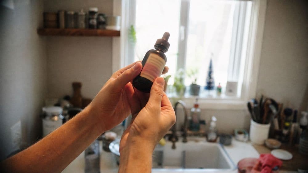 A person tilting a dropper bottle toward a kitchen window to read the small print on the back label, the text too small and angled to be legible.