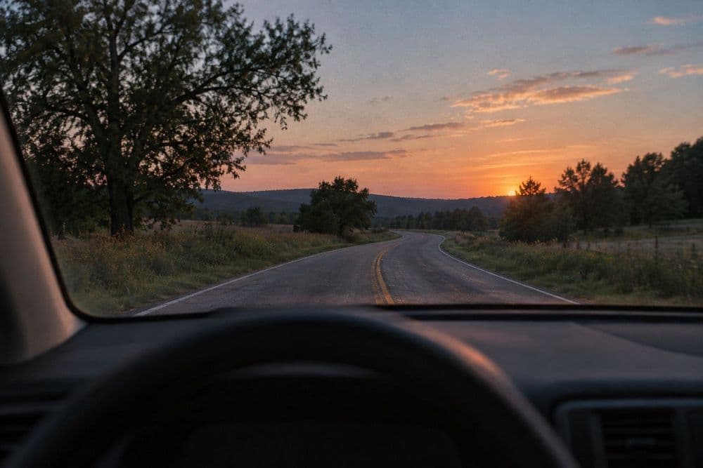 A quiet two-lane road at dusk seen through a car windshield, the dashboard and steering wheel soft in the foreground, with no people visible.