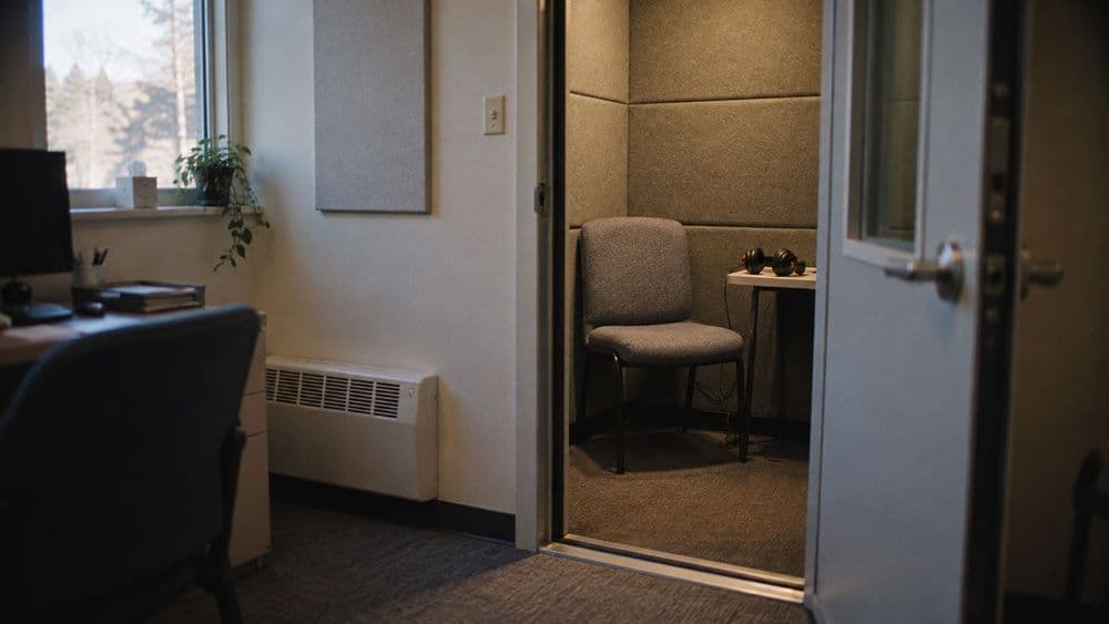 An empty audiology booth with a padded chair, a pair of over-ear test headphones resting on a small table, and soft daylight through a small window.
