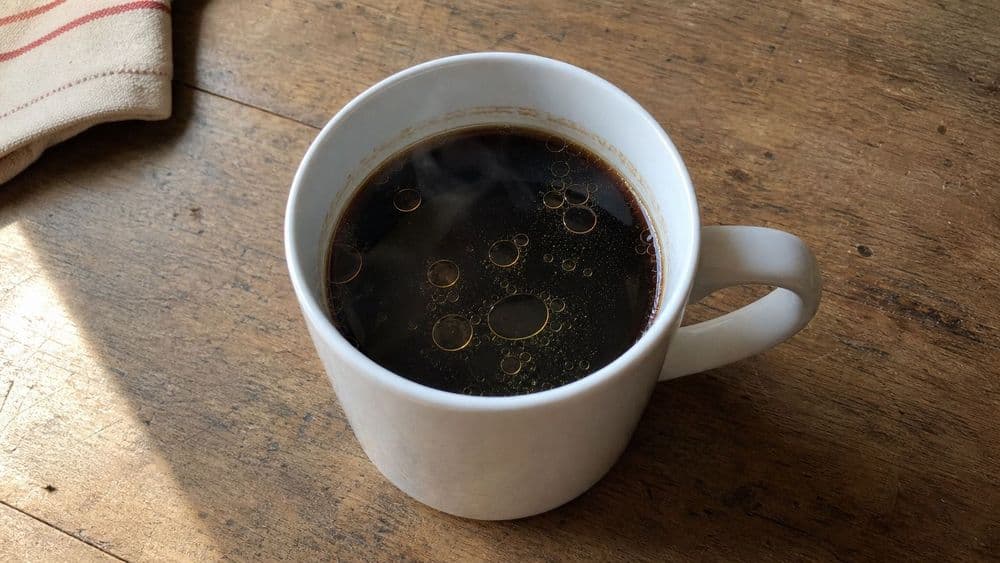 Looking down into a plain white mug of black coffee on a worn wooden table, a thin film of golden oil broken into separated round islands across the surface and a faint oily ring clinging to the inside of the rim.