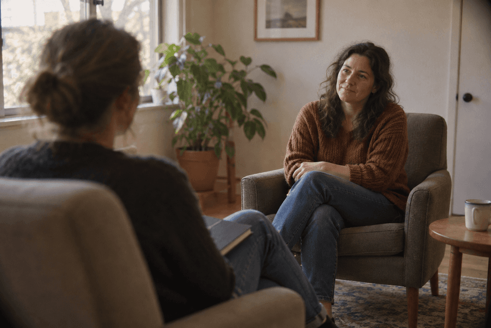 Two adults talking in a quiet, sunlit counseling room, one seen over the shoulder with a closed notebook on their knee, the other listening attentively.